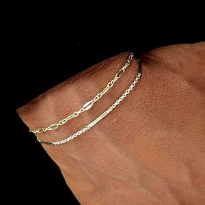 May include: A close-up of three delicate chain bracelets. The top bracelet is gold-toned with oval links, the middle one is a silver-toned chain, and the bottom one is a silver-toned chain. The bracelets are worn on a wrist against a dark background.