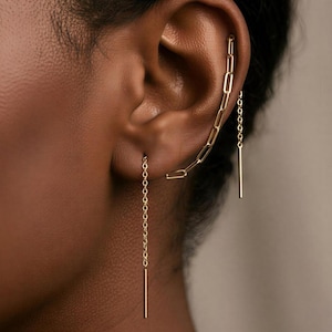 Pode incluir: Um brinco ear crawler dourado com uma série de elos retangulares alongados que seguem a curva da orelha, ligado a uma corrente delicada que pende do lóbulo da orelha, terminando num pendente fino de barra vertical reta.