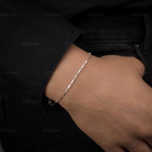 May include: A silver bracelet with a rectangular link chain is worn on a person's wrist. The bracelet is simple and elegant, with a delicate design. The background is a dark color, which makes the bracelet stand out.