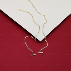 Op de afbeelding: Een paar goudkleurige oorhangers. Elke oorbel heeft een delicate ketting met een kleine staaf aan het uiteinde. De oorbellen worden gepresenteerd op een wit oppervlak tegen een rode achtergrond.