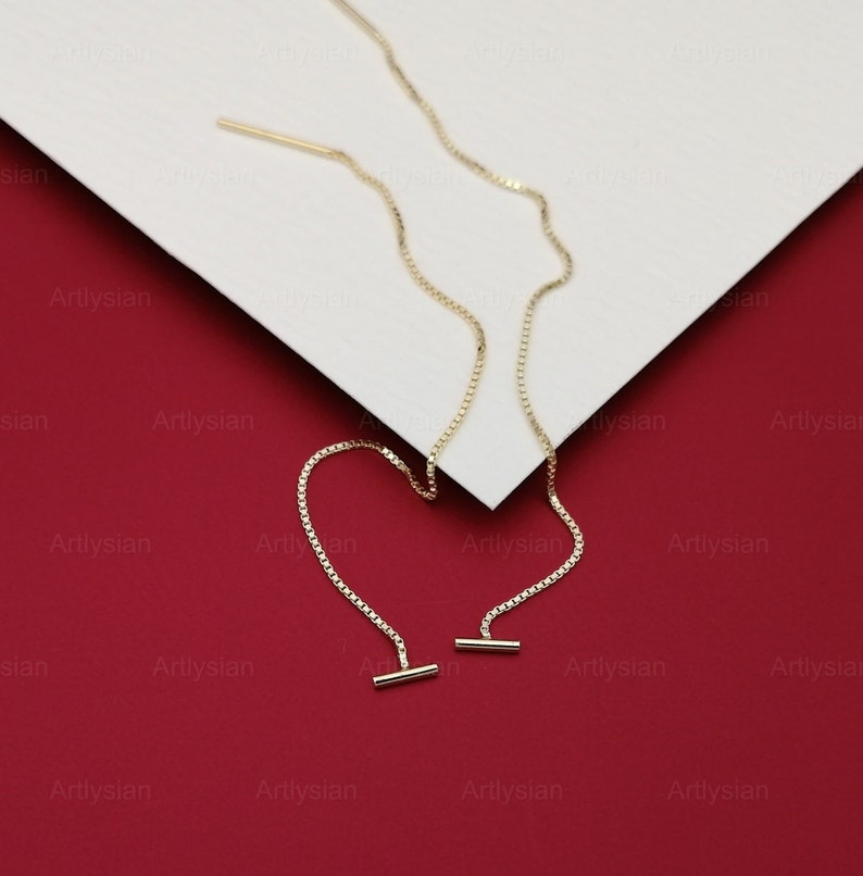 Op de afbeelding: Een paar goudkleurige oorhangers. Elke oorbel heeft een delicate ketting met een kleine staaf aan het uiteinde. De oorbellen worden gepresenteerd op een wit oppervlak tegen een rode achtergrond.
