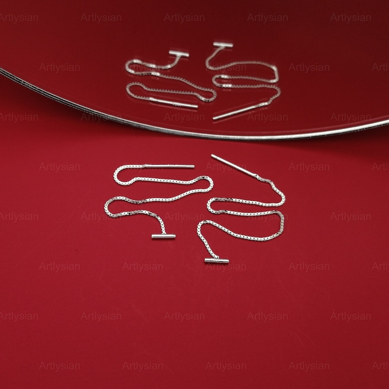 Op de afbeelding: Zilveren oorhangers met een T-bar design en een delicate ketting. De oorbellen worden gepresenteerd op een rode ondergrond met een zilveren rand. Een eenvoudig en elegant accessoire.