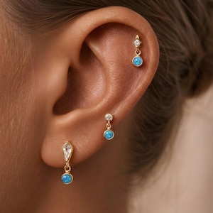 Puede incluir: Primer plano de tres pendientes dorados con piedras azules similares al ópalo y cristales transparentes. Uno es un pendiente de botón con un solo cristal y ópalo. Los otros dos son colgantes, con un diseño de cristal y ópalo.