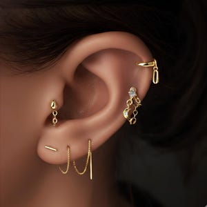 Könnte beinhalten: Nahaufnahme eines Ohrs mit mehreren goldenen Ohrringen. Die Ohrringe umfassen einen kleinen Tropfen mit Kette, einen Stecker, Kettenohrringe und ein Monddesign mit einem kleinen Edelstein. Der Schmuck ist goldfarben.