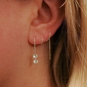 May include: A close-up of a person's ear with multiple earrings. The earrings are silver and feature a small chain with a clear crystal at the end. The earrings are worn in the helix and lobe piercings.