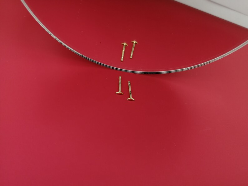 May include: Two pairs of gold stud earrings on a red background. The earrings are small and have a simple design.