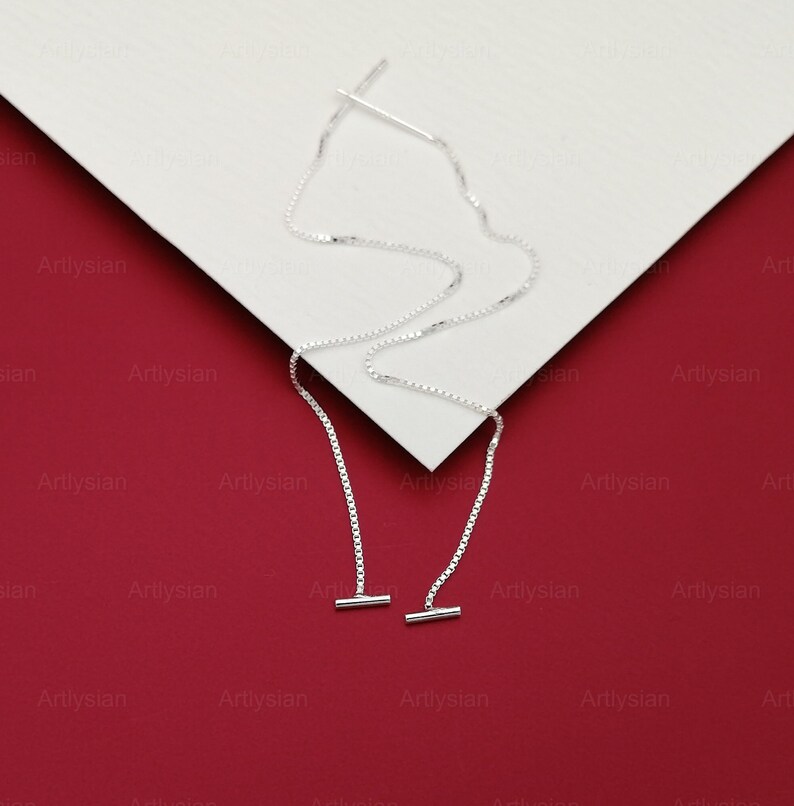 Op de afbeelding: Een paar zilveren oorhangers. Elke oorbel heeft een fijne ketting die aan een rechte staaf hangt, met een kleine staaf aan het uiteinde. De oorbellen worden getoond op een wit oppervlak tegen een rode achtergrond.
