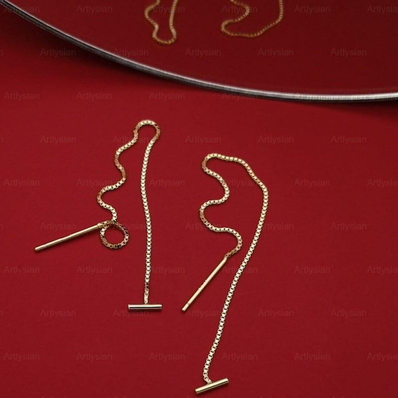 Op de afbeelding: Goudkleurige oorhangers met een T-bar en ketting ontwerp. De oorbellen worden gepresenteerd op een rode ondergrond, met het woord "Artlysian" herhaald op de achtergrond.