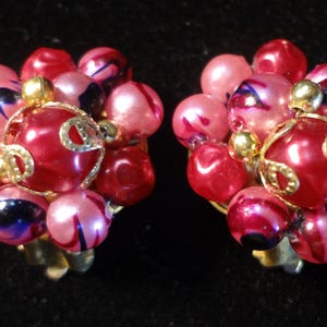 May include: A pair of vintage clip-on earrings. Each earring features a cluster of pink, red, and purple beads. The center bead is a larger, glossy red with gold-tone filigree. The earrings have gold-tone clips.