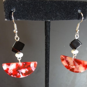 May include: A pair of dangle earrings featuring a silver-toned hook, a small black cube bead, a tiny white spacer bead, and a semi-circular pendant with a marbled red and white pattern. The earrings hang from a black display stand.