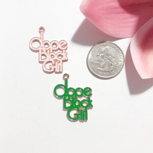 May include: Two metal charms with the words "Dope Black Girl" in pink and green enamel. The charms are on a white background.