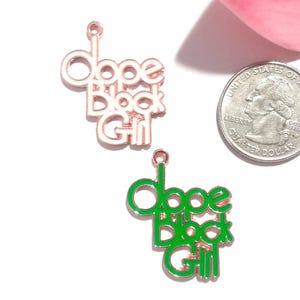 May include: Two metal charms with a rose gold finish, each featuring the text "Dope Black Girl" in a stacked design. One charm has white enamel fill, and the other has green enamel fill. A US quarter coin is shown for scale next to the charms.
