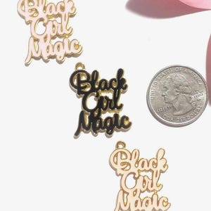 May include: Three metal charms featuring the text Black Girl Magic in a script font. Two charms are light pink with gold-tone edges, and one is black with gold-tone edges. A United States quarter is included for scale, measuring 0.955 inches in diameter.