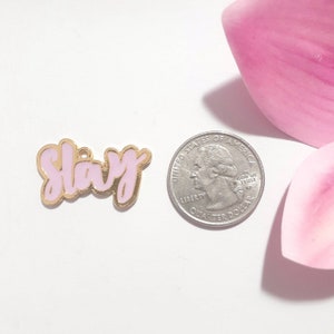 May include: A pink and gold enamel pin with the word "Slay" in a stylized font. The pin is about the size of a US quarter.