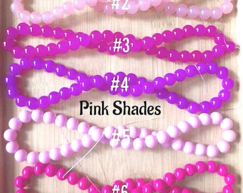 May include: Seven strands of pink beads in different shades, labeled #1 through #7. The beads are arranged in a row on a wooden surface. The text "Pink Shades" is written below the beads.