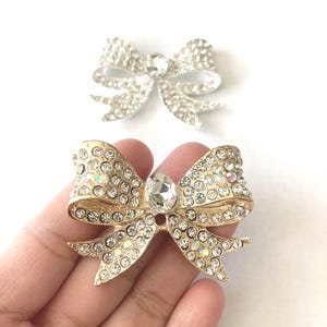 May include: Two rhinestone bow brooches, one gold and one silver. Both bows are adorned with clear rhinestones and have a large, clear rhinestone in the center.