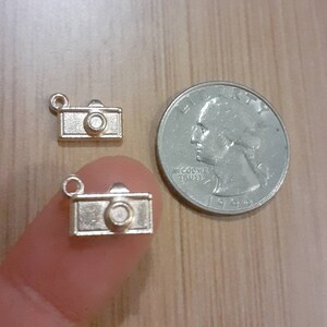 May include: Two gold-toned camera charms, one on a finger and one on a wooden surface, next to a US quarter. The quarter is dated 1994 and has the words "In God We Trust" on it.