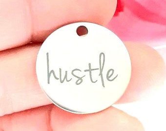 May include: A silver round charm with the word "hustle" engraved in a cursive font.