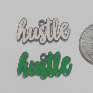 May include: Two metal charms in cursive script reading hustle, one with white enamel and one with green enamel, positioned next to a United States quarter for scale. Each charm features a small circular loop at the top for attachment.
