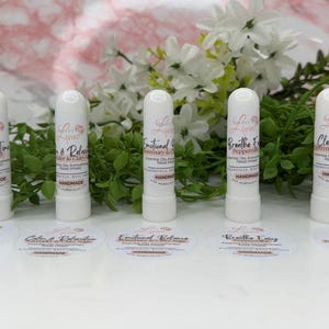 May include: Five white roll-on essential oil inhalers with labels. The labels are white with black text and pink accents. The labels read "Happy Time", "Calm & Relax", "Emotional Balance", "Breathe Easy", and "Clear Focus".