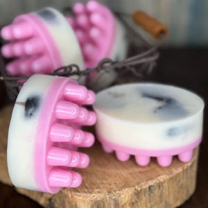 May include: Pink and white massage soap bars with rounded massage heads. The soap bars are displayed on a wooden surface and in a wire basket. The soap bars have a white center with pink massage heads and dark gray accents.