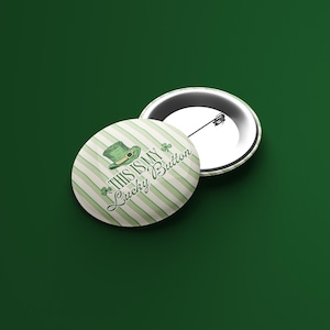 Puede incluir: Dos insignias redondas con temática del Día de San Patricio. Una insignia muestra un sombrero verde de duende y el texto "THIS IS MY Lucky Button" sobre un fondo a rayas. La otra insignia está boca abajo, mostrando el alfiler.