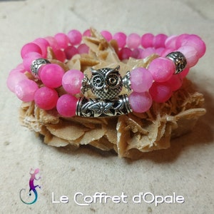 Peut inclure: Deux bracelets en agate rose avec un charme hibou argenté. Les bracelets sont empilés l'un sur l'autre et reposent sur une surface brun clair.