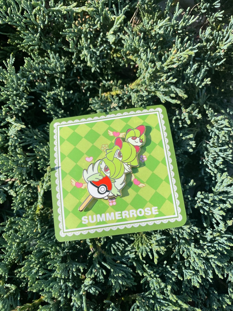 May include: A metal enamel pin featuring three stacked green and white humanoid figures with red accents, one holding a red and white ball, attached to a square green checkered backing card with the white text SUMMERROSE printed at the bottom.