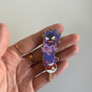 May include: A metal enamel pin featuring a vertical stack of three purple ghost-like creatures, including Gastly, Haunter, and Gengar, balanced on top of a red and white Pok&eacute; Ball with a small gold-colored stick extending from the bottom.
