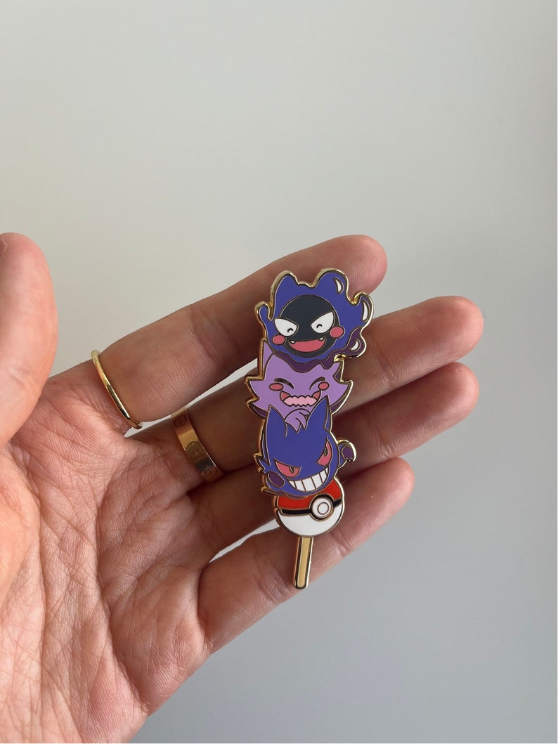 May include: A metal enamel pin featuring a vertical stack of three purple ghost-like creatures, including Gastly, Haunter, and Gengar, balanced on top of a red and white Pok&eacute; Ball with a small gold-colored stick extending from the bottom.
