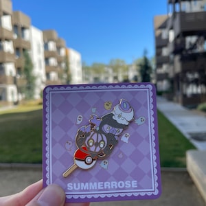 May include: A metal enamel pin featuring a stacked design of three ghost-type creatures atop a red and white Pok&eacute; Ball on a stick. The pin is displayed on a square purple checkered backing card with the text SUMMERROSE printed at the bottom.