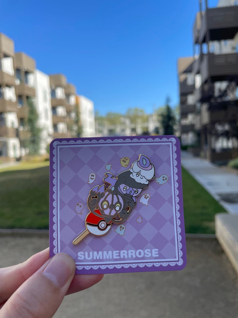 May include: A metal enamel pin featuring a stacked design of three ghost-type creatures atop a red and white Pok&eacute; Ball on a stick. The pin is displayed on a square purple checkered backing card with the text SUMMERROSE printed at the bottom.