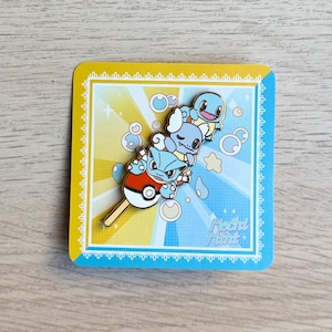May include: A colorful enamel pin featuring three cartoon characters on a popsicle stick. The pin is set against a yellow and blue background with bubbles and stars. The text "Flachi Mint" is at the bottom.