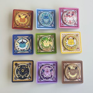 May include: Nine square-shaped pins featuring different Pokemon characters. Each pin has a stained glass design with a gold border and the Pokemon's name printed below. The Pokemon featured are: Flareon, Glaceon, Sylveon, Vaporeon, Leafeon, Jolteon, Umbreon, Espeon, and  Flareon.
