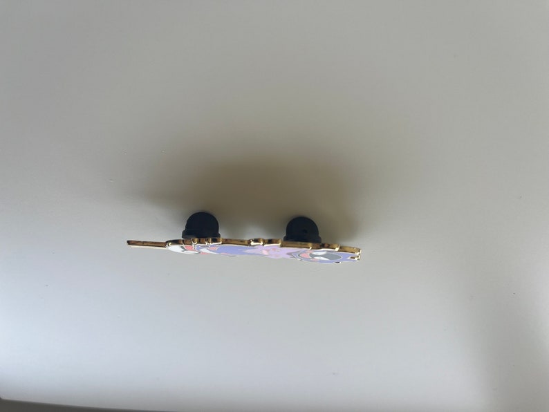 May include: A metal enamel pin viewed from the side, showing the thin profile of the decorative piece. Two black rubber clutch backings are attached to the metal posts on the reverse side of the pin, which features gold-toned metal edges.