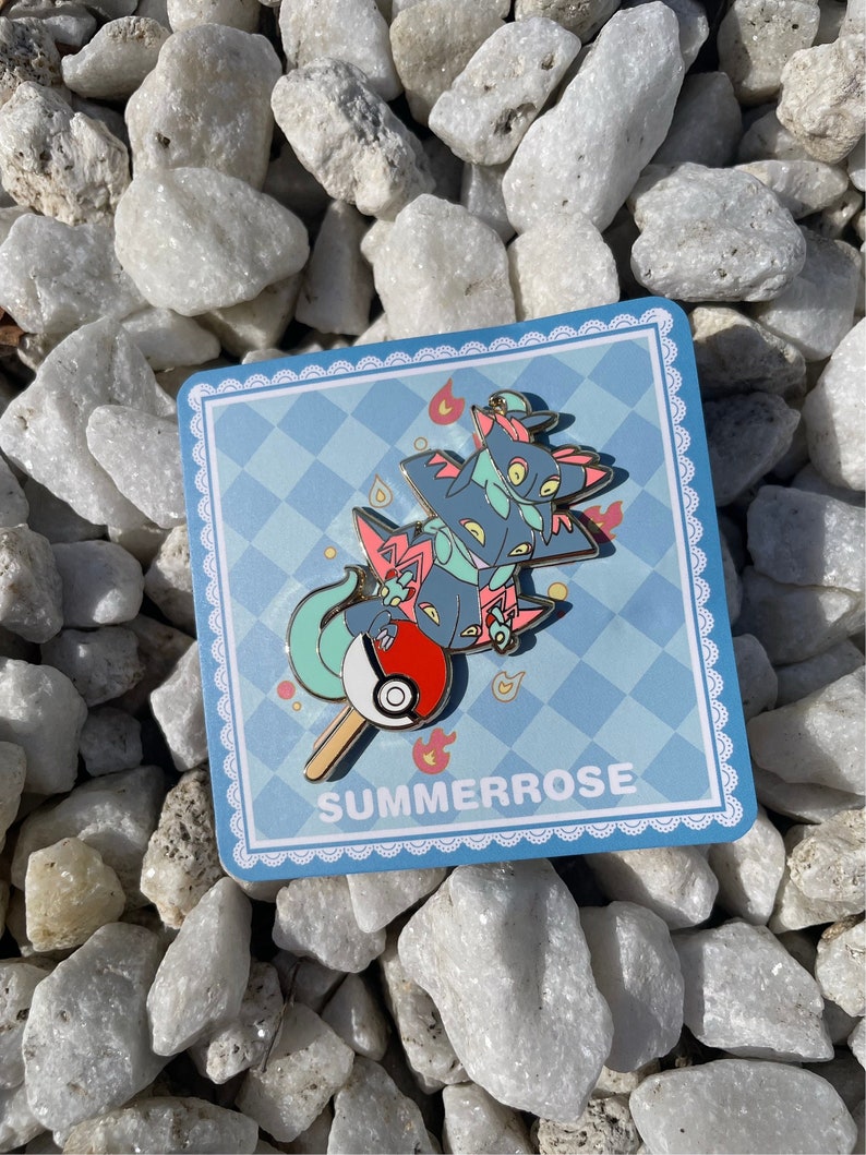 May include: A metal enamel pin featuring a stack of three dark blue and teal dragon-like creatures balanced on a red and white ball, all on a stick. The pin is attached to a square blue and white checkered backing card with the text SUMMERROSE.