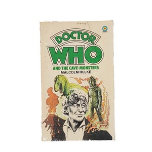 May include: A vintage paperback book cover featuring the Doctor Who logo in green and white. The cover depicts the Fourth Doctor, a dinosaur, and a green alien creature. The title reads "Doctor Who and the Cave-Monsters" by Malcolm Hulke.