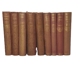 Pode incluir: Uma coleção de nove livros antigos de Charles Dickens com capas de tecido castanho e letras douradas nas lombadas. Os títulos incluem The Pickwick Papers, Dombey and Son, Barnaby Rudge, David Copperfield e Great Expectations.