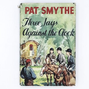 Puede incluir: Una portada de libro vintage con el título "Pat Smythe Three Jays Against the Clock". La portada presenta una ilustración de tres niños a caballo frente a una caravana tirada por caballos.