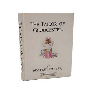 Op de afbeelding: Een vintage boekomslag voor "De kleermaker van Gloucester" van Beatrix Potter. De omslag toont een bruine muis die op een spoel garen zit, een stuk stof vasthoudt en naar een naaipatroon kijkt.