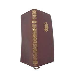 May include: A maroon hardcover book titled Captains Courageous by Rudyard Kipling. The spine features gold-colored botanical illustrations and text. A circular gold emblem is visible on the front cover. The book has a small ribbon bookmark at the base.