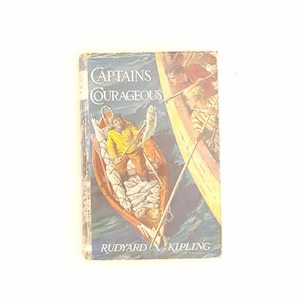 May include: A hardcover book titled Captains Courageous by Rudyard Kipling. The cover features a color illustration of a person in a yellow jacket holding a large fish in a small boat, with another person in a boat nearby on blue water.