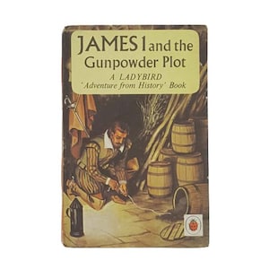 Op de afbeelding: Een Ladybird 'Adventure from History' boekomslag met een illustratie van een man in een bruin-wit gestreepte outfit die een brandende lont vasthoudt in de buurt van een groep vaten. De titel van het boek is "JAMES 1 and the Gunpowder Plot".