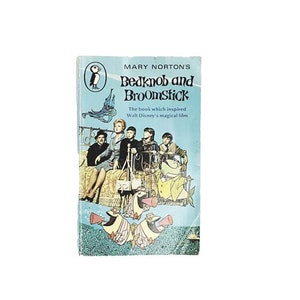 May include: A vintage paperback book cover featuring a colorful illustration of a group of people riding a bed on a broomstick. The book title is "Bedknob and Broomstick" by Mary Norton. The text below the title reads "The book which inspired Walt Disney's magical film."