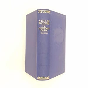 Pode incluir: Um livro de capa dura azul com texto dourado em relevo na lombada que diz A TALE OF TWO CITIES A CHRISTMAS CAROL DICKENS. A capa tem uma borda decorativa sutil em relevo e a lombada inclui ornamentos dourados na parte superior e inferior.