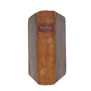 May include: An antique book with a leather spine and cover. The spine is a warm brown color with the title "Byron" in gold lettering on a red label. The book's edges are a muted gray.
