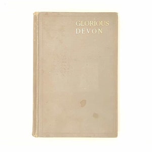 Może przedstawiać: Książka w kolorze beżowym z tytułem "Glorious Devon" nadrukowanym złotymi literami na okładce.