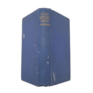 Może przedstawiać: Vintage niebieska książka zatytułowana "What Katy Did Next" autorstwa Coolidge'a. Książka ma złote litery i nosi ślady użytkowania, z widocznymi zadrapaniami i śladami na okładce. Grzbiet ma wytłoczone detale.