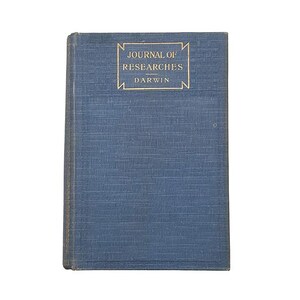 Op de afbeelding: Een blauw vintage boek met de titel "Journal of Researches Darwin" in gouden letters. De titel staat in een gouden rechthoekige lijst. Het boek heeft een zichtbare textuur. De achtergrond is wit.