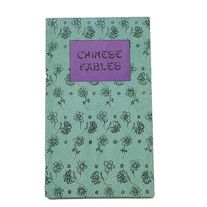 Puede incluir: Un libro rectangular titulado CHINESE FABLES con una cubierta de color verde claro que presenta un patrón repetitivo de pequeñas flores y puntos dibujados en negro. Una etiqueta rectangular morada está centrada en la portada.